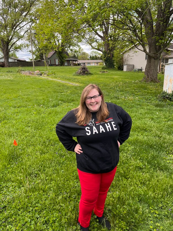 Derin is wearing a Ball State SAAHE sweatshirt, standing outside, smiling